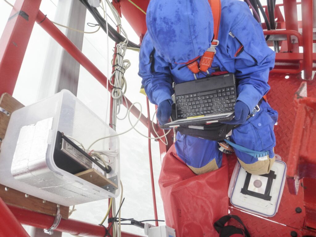 person in blue snowsuit and climbing gear sitting on the tower with notebook on his lap reading a device in cold and wet weather in winter